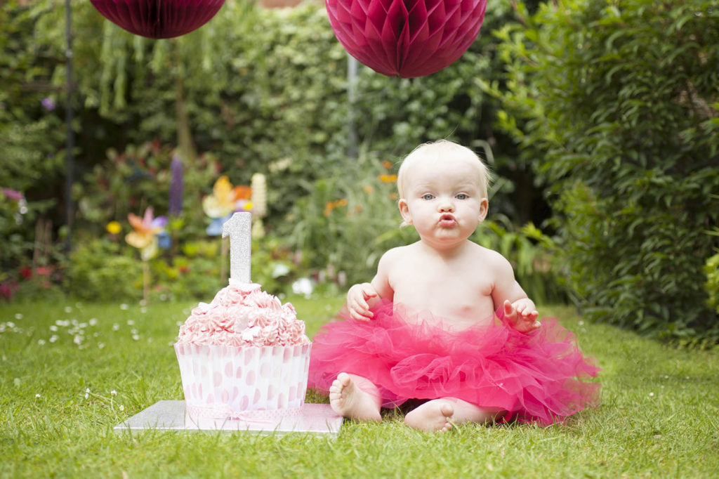 Cheshire cake smash photography of a one year old baby in a pink tutu next to a birthday cake with a number one on it.