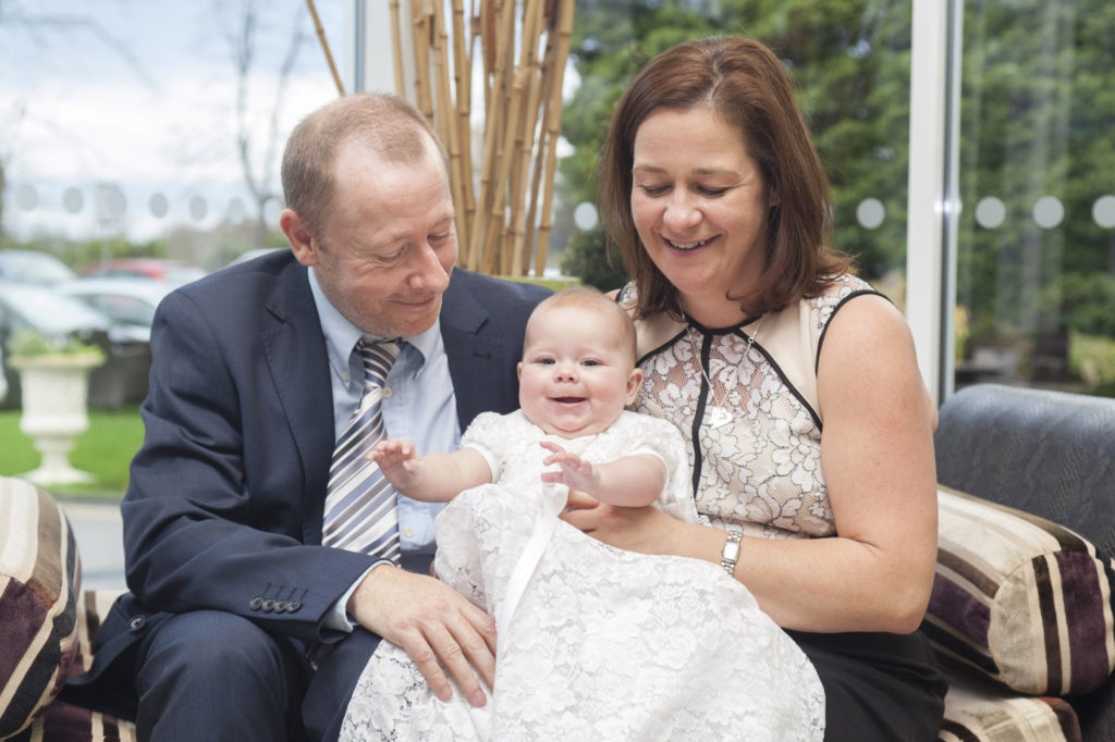 Family photography of a Mum and Dad holding a baby wearing a christening gown.