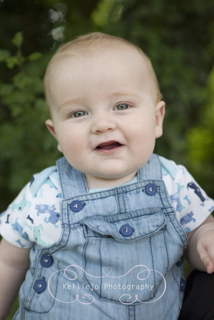 Children's photography of a young baby dressed in denim dungarees smiling directly at the camera.